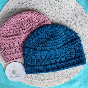 Puede incluir: Dos gorros de ganchillo hechos a mano en rosa y azul verdoso, sobre un mantel individual de encaje blanco. Los gorros tienen un patr&oacute;n de puntada texturizada. Tambi&eacute;n hay una peque&ntilde;a etiqueta redonda con una ilustraci&oacute;n de b&uacute;ho y texto.