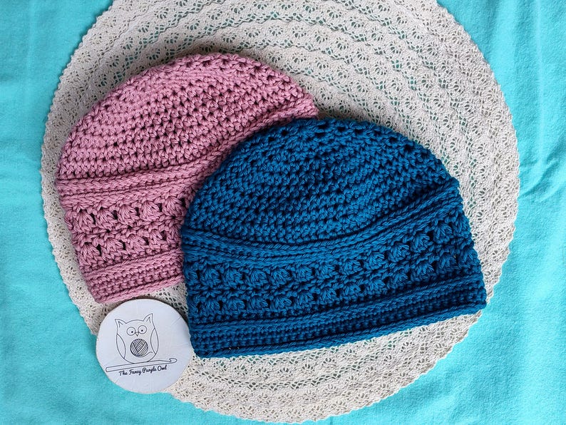 Puede incluir: Dos gorros de ganchillo hechos a mano en rosa y azul verdoso, sobre un mantel individual de encaje blanco. Los gorros tienen un patr&oacute;n de puntada texturizada. Tambi&eacute;n hay una peque&ntilde;a etiqueta redonda con una ilustraci&oacute;n de b&uacute;ho y texto.