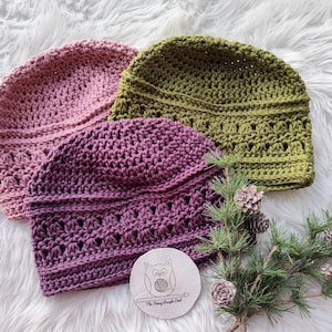 Puede incluir: Tres gorros de ganchillo hechos a mano en tonos rosa, morado y verde oliva se exhiben sobre una superficie blanca y esponjosa. Los gorros tienen un patr&oacute;n de puntada texturizada. Tambi&eacute;n hay un peque&ntilde;o disco de madera con un logotipo de b&uacute;ho y ramitas de pino con pi&ntilde;as.