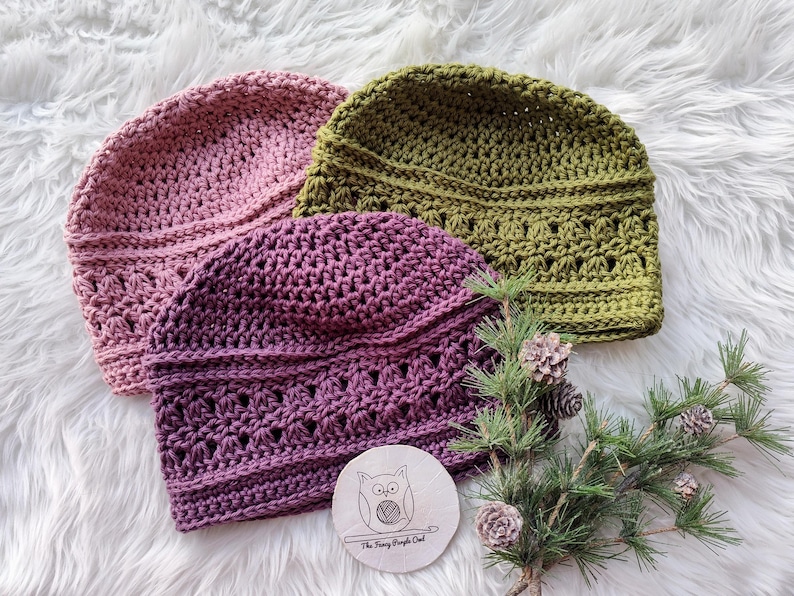 Puede incluir: Tres gorros de ganchillo hechos a mano en tonos rosa, morado y verde oliva se exhiben sobre una superficie blanca y esponjosa. Los gorros tienen un patr&oacute;n de puntada texturizada. Tambi&eacute;n hay un peque&ntilde;o disco de madera con un logotipo de b&uacute;ho y ramitas de pino con pi&ntilde;as.
