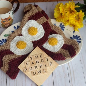 Puede incluir: Una bufanda de ganchillo con un patr&oacute;n ondulado en color canela y granate oscuro, decorada con tres apliques de huevos fritos en blanco y amarillo. Un cuadrado de madera muestra el texto THE FANCY PURPLE OWL en letras negras sobre el tejido.