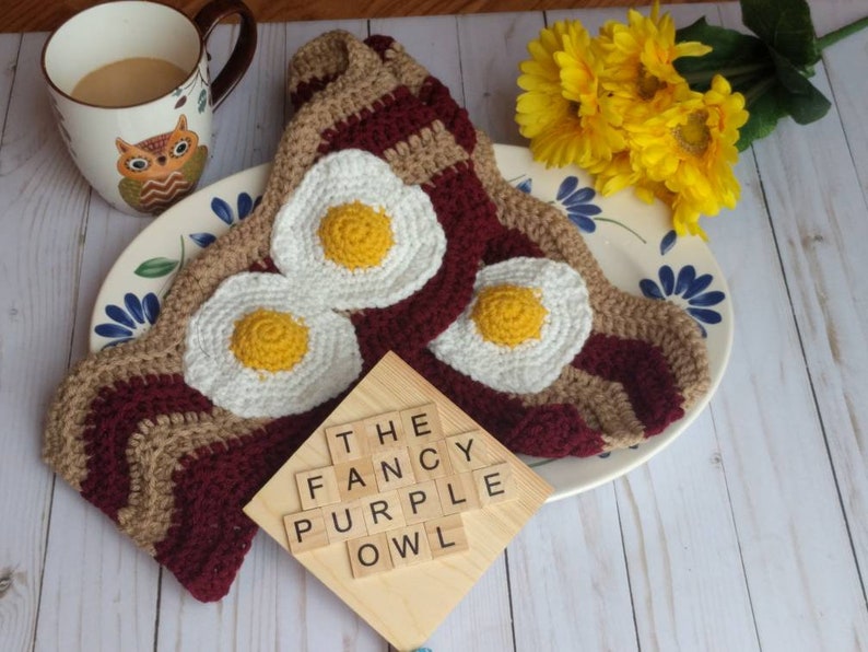 Puede incluir: Una bufanda de ganchillo con un patr&oacute;n ondulado en color canela y granate oscuro, decorada con tres apliques de huevos fritos en blanco y amarillo. Un cuadrado de madera muestra el texto THE FANCY PURPLE OWL en letras negras sobre el tejido.