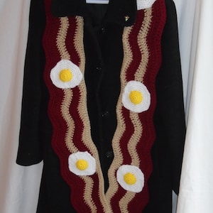 Puede incluir: Una bufanda larga de ganchillo con un patr&oacute;n ondulado en rojo oscuro y beige, que recuerda a tiras de tocino, decorada con varios motivos circulares de ganchillo en forma de huevos fritos blancos y amarillos.