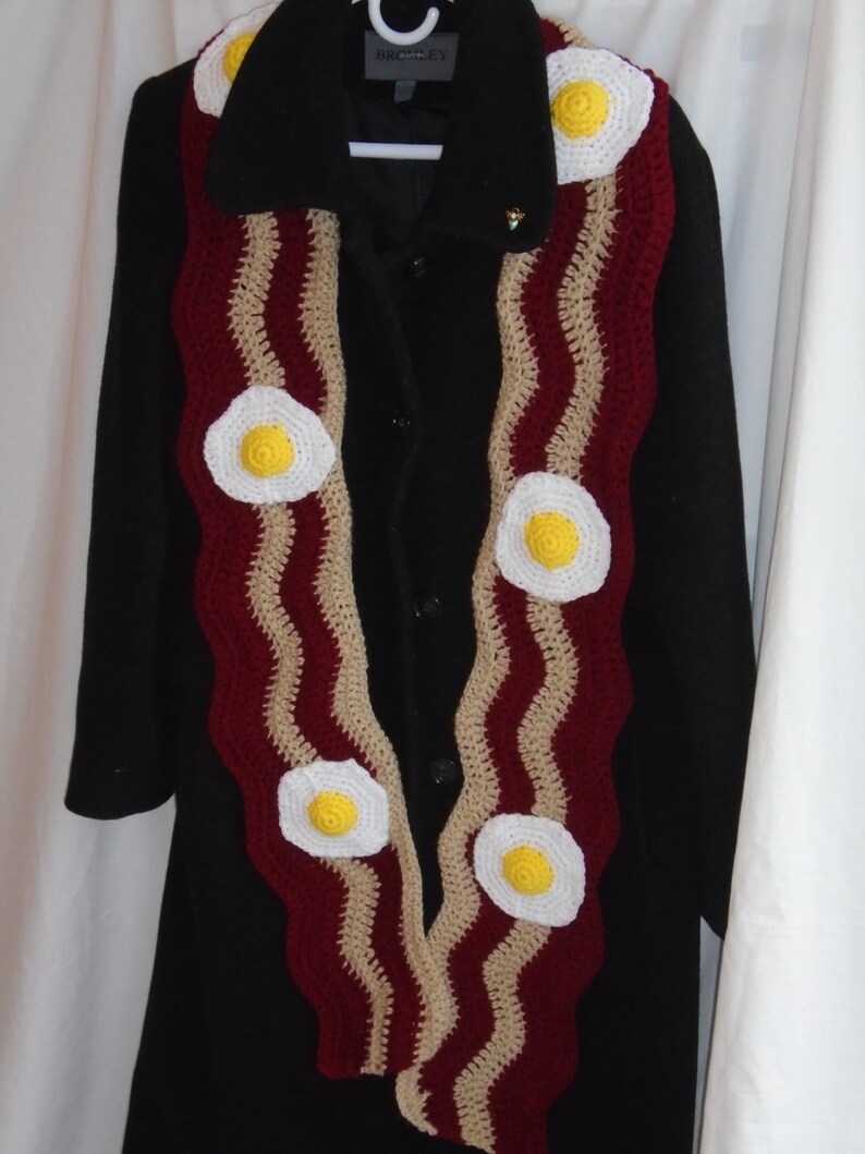 Puede incluir: Una bufanda larga de ganchillo con un patr&oacute;n ondulado en rojo oscuro y beige, que recuerda a tiras de tocino, decorada con varios motivos circulares de ganchillo en forma de huevos fritos blancos y amarillos.