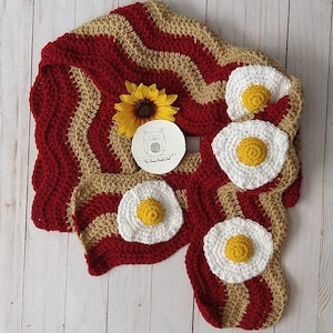 Puede incluir: Una bufanda de ganchillo con un patr&oacute;n ondulado de rayas rojo oscuro y beige que recuerda al tocino, decorada con cuatro aplicaciones circulares de ganchillo en forma de huevo frito blanco y amarillo. Incluye una peque&ntilde;a etiqueta redonda con un logo de b&uacute;ho.