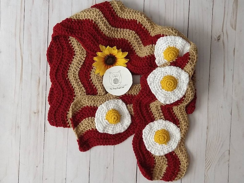 Puede incluir: Una bufanda de ganchillo con un patr&oacute;n ondulado de rayas rojo oscuro y beige que recuerda al tocino, decorada con cuatro aplicaciones circulares de ganchillo en forma de huevo frito blanco y amarillo. Incluye una peque&ntilde;a etiqueta redonda con un logo de b&uacute;ho.
