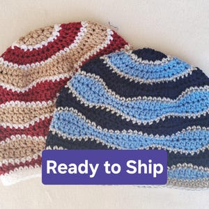May include: Two handmade crochet beanies. One beanie features alternating stripes of red, white, and tan. The other beanie has blue and navy stripes with gray accents. The beanies are ready to ship.