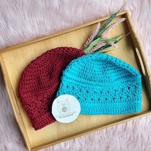 Puede incluir: Dos gorros de ganchillo, burdeos y azul turquesa, descansan sobre una bandeja de madera. El gorro burdeos tiene un patr&oacute;n liso, mientras que el turquesa presenta un dise&ntilde;o calado decorativo. Tambi&eacute;n hay una peque&ntilde;a etiqueta redonda con una ilustraci&oacute;n de un b&uacute;ho.