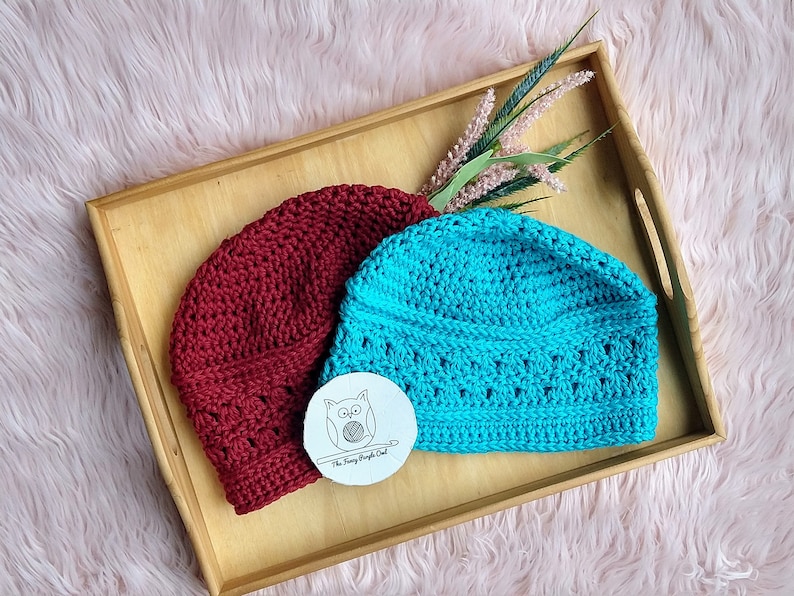 Puede incluir: Dos gorros de ganchillo, burdeos y azul turquesa, descansan sobre una bandeja de madera. El gorro burdeos tiene un patr&oacute;n liso, mientras que el turquesa presenta un dise&ntilde;o calado decorativo. Tambi&eacute;n hay una peque&ntilde;a etiqueta redonda con una ilustraci&oacute;n de un b&uacute;ho.