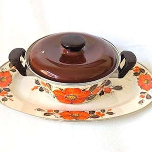 May include: A vintage casserole dish with a matching oval serving platter. The dish has a brown lid and handles, and the set features a floral design with orange flowers and brown leaves on a cream background.