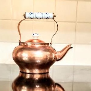 May include: A copper teapot with a white porcelain handle and lid. The teapot has a rounded body and a spout on the side.