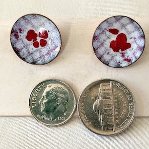 May include: Two small, round, copper earrings with white enamel and red enamel accents. The earrings are about the size of a dime.