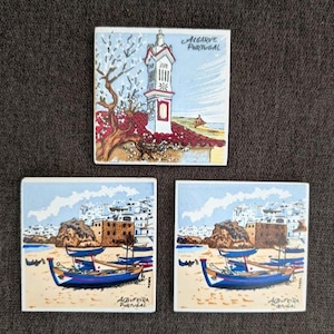 May include: Three square ceramic tiles with colorful illustrations of Algarve, Portugal. One tile depicts a tower with the text "Algarve Portugal". The other two tiles show boats on a beach with the text "Albufeira Portugal".