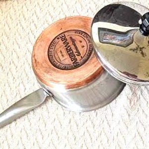 May include: A Farberware Advantage stainless steel saucepan with a copper impact base and a matching stainless steel lid. The bottom of the pan features a circular stamp with the brand name, 18/10 stainless steel, and copper impact base text.