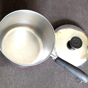 May include: A small, stainless steel saucepan with a black handle and a matching lid. The lid has a black knob. The pan is empty and sits on a gray surface. The handle has a loop for hanging.