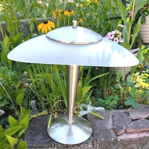 May include: A table lamp featuring a brushed metal conical base and a circular stand. The lamp has a wide, frosted glass shade shaped like a shallow dome, topped with a small, polished metal cap at the center of the upper surface.