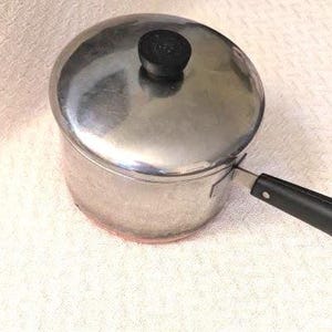 May include: A stainless steel saucepan with a polished finish, featuring a matching lid with a black circular knob on top. The pan includes a long, black heat-resistant handle attached to the side, providing a functional grip for cooking tasks.