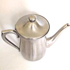 May include: A silver-colored teapot with a lid and a curved spout. The teapot has a handle on the side and a small knob on the lid. The teapot is made of metal and has a shiny finish. The teapot is designed for serving tea.