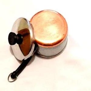 May include: A small round copper-colored pot with a silver-toned lid. The lid features a black knob and a long black handle with a metal loop at the end. The pot has a metallic finish and is positioned next to the lid on a plain white background.
