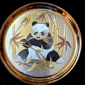 May include: A circular decorative plate featuring a metallic gold and silver finish. The center depicts a panda bear sitting and eating bamboo stalks. The design is rendered in fine, etched lines with a polished gold-toned rim and a black border.