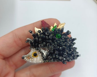 Puede incluir: Un broche de erizo de cuentas negras y blancas con una baya roja y hojas verdes en la parte superior. El broche está hecho a mano y tiene un ojo de cristal.