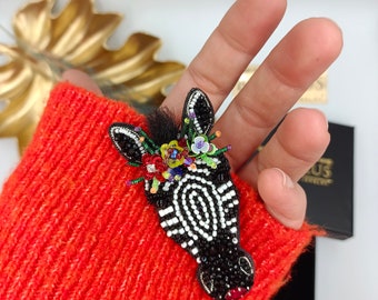 Peut inclure: Une broche en forme de zèbre faite de perles de rocaille noires et blanches, ornée d'une couronne florale de sequins et de perles colorées entre les oreilles. La tête de zèbre présente une touffe de poils noirs et un accent rouge à la bouche.