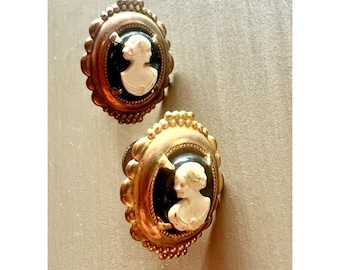 Könnte beinhalten: Zwei ovale, goldfarbene antike Ohrringe mit einem Cameo-Design. Jeder Ohrring hat einen schwarzen Hintergrund mit einem weißen Relief eines Frauenprofils. Die Ohrringe haben einen gewellten Rand und einen detaillierten Rahmen.