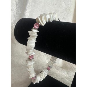 May include: A white shell bracelet with pink beads and silver accents. The bracelet is made of irregular, flat shell pieces strung together. The pink beads are interspersed throughout the bracelet, adding a pop of color. The bracelet is displayed on a black stand.