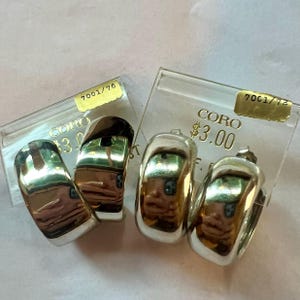 May include: Two pairs of silver hoop earrings with a smooth, rounded finish. The earrings are packaged in clear plastic boxes with price tags that read "Coro $3.00" and "Coro $3.00".