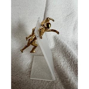 May include: A gold-toned metal sculpture featuring two stylized human figures in motion, attached to a clear, angled acrylic stand. The figures are depicted in a running or leaping pose, with one figure positioned slightly behind and above the other.