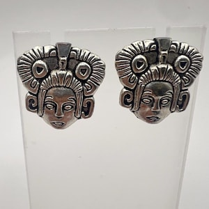 May include: A pair of silver-toned stud earrings. Each earring features a detailed face design with an elaborate headdress. The earrings are displayed on a clear, angled stand against a white background. The design has an ancient, possibly Mayan, aesthetic.