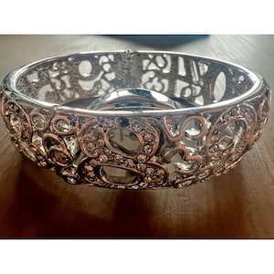 May include: Silver-toned hinged bangle bracelet with an openwork design. The bracelet features a repeating pattern of hearts and swirls, embellished with clear crystals. The bracelet is resting on a wooden surface.