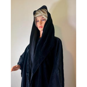 May include: A black velvet hooded cloak. The cloak has a wide hood and drapes over the shoulders. The fabric appears soft and luxurious. The cloak is a solid black color and is displayed on a mannequin.