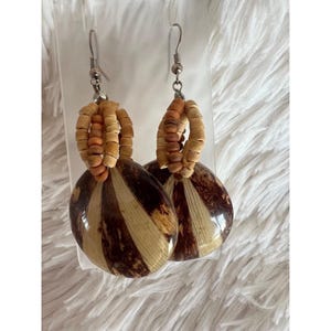 May include: A pair of dangle earrings featuring silver-toned metal hooks, a cluster of small round wooden beads in shades of tan and brown, and a large, flat, circular pendant with alternating vertical stripes of dark brown and light beige.
