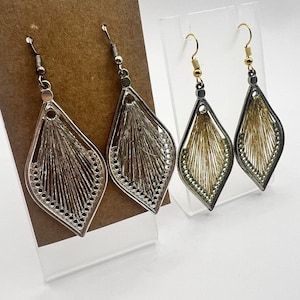 Puede incluir: Dos pares de pendientes colgantes. Un par es plateado con un interior tejido de color marrón claro. El otro par es dorado con un interior tejido de color marrón claro. Ambos pares tienen forma de lágrima con un borde decorativo.