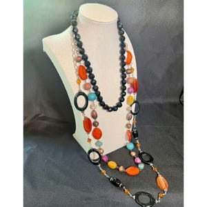 May include: A multi-strand necklace with a variety of colourful beads and accents. The necklace features black, orange, blue, and yellow beads, along with black oval-shaped pendants. The necklace is displayed on a white mannequin.