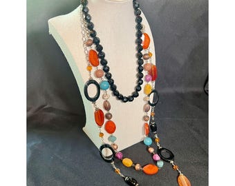 May include: A multi-strand necklace with a variety of colorful beads and accents. The necklace features black, orange, blue, and yellow beads, along with black oval-shaped pendants. The necklace is displayed on a white mannequin.