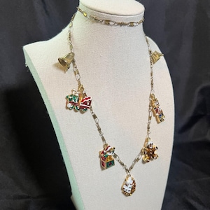 Peut inclure: Un collier doré sur le thème de Noël avec divers breloques, dont des cloches, un train, un bonhomme en pain d'épices et des boîtes cadeaux. Les breloques sont détaillées avec de l'émail rouge, vert et blanc. Le collier est présenté sur un mannequin.