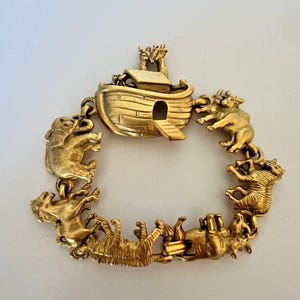 Puede incluir: Un brazalete dorado con una representación detallada del Arca de Noé con animales que la rodean. El arca tiene una pequeña abertura y una rampa. Los animales incluyen una cebra, un elefante, un león y una vaca.