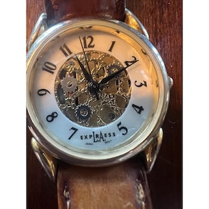 May include: A gold-toned Expiraess watch with a brown leather band. The watch face has a white background with black numerals and visible gears. The watch reads "Expiraess Japan Movt."
