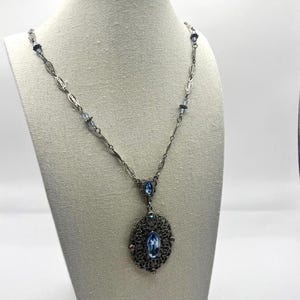 May include: A silver-toned necklace with a filigree pendant and light blue oval-shaped gemstone. The necklace features a delicate chain with small blue accents and is displayed on a mannequin.