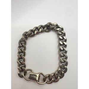 Puede incluir: Una pulsera de cadena de eslabones plateada con un cierre seguro. La pulsera presenta eslabones gruesos y redondeados y un cierre grabado con la palabra "LISNER". La joya se muestra sobre un fondo blanco.
