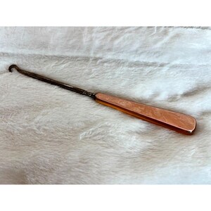 May include: A vintage copper-toned shoe horn with a curved hook at one end and a flat, rectangular handle. The handle has a warm, reddish-brown hue, and the metal has a slightly aged appearance.