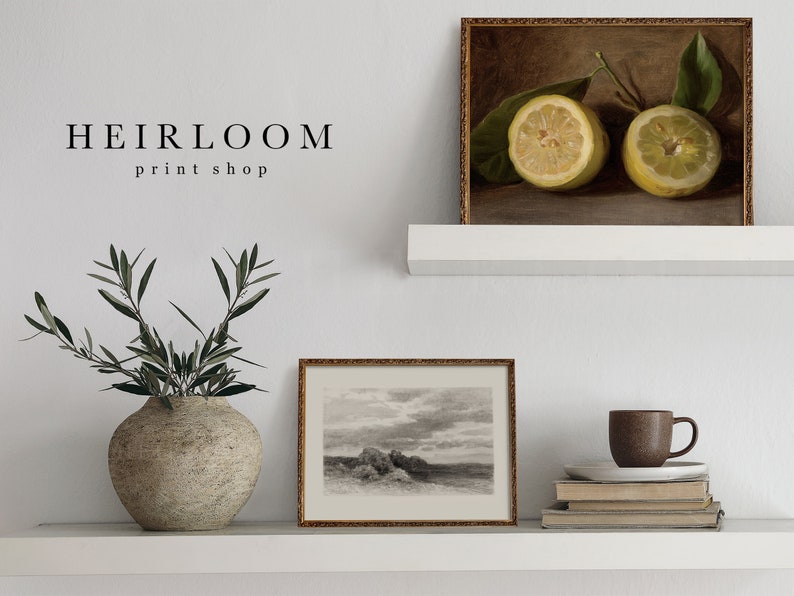 May include: A still life print of two lemon halves with green leaves in a gold frame. Below, a black and white landscape print and a vase with olive branches sit on a white shelf. The text "HEIRLOOM print shop" is above the vase.