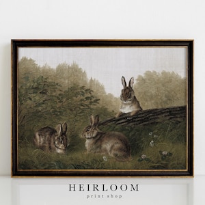 May include: A framed vintage print featuring three rabbits in a natural setting. The artwork depicts the rabbits in a grassy area near a log and trees, with a muted color palette of greens, browns, and grays. The text "HEIRLOOM print shop" is at the bottom.