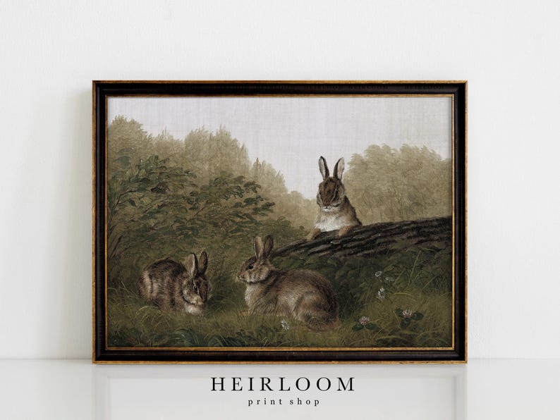 May include: A framed vintage print featuring three rabbits in a natural setting. The artwork depicts the rabbits in a grassy area near a log and trees, with a muted color palette of greens, browns, and grays. The text "HEIRLOOM print shop" is at the bottom.