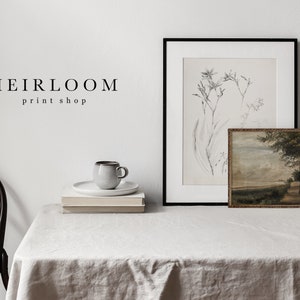 May include: A white table with a white tablecloth, a black framed print of a botanical illustration, a small framed landscape painting, a white ceramic mug, and two stacks of books. The text "HEIRLOOM print shop" is visible on the wall.