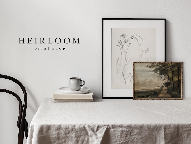 May include: A white table with a white tablecloth, a black framed print of a botanical illustration, a small framed landscape painting, a white ceramic mug, and two stacks of books. The text "HEIRLOOM print shop" is visible on the wall.