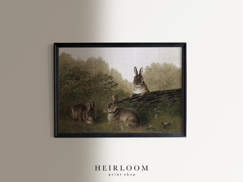 May include: Framed print featuring three rabbits in a naturalistic setting. The artwork depicts the rabbits in a grassy area with a log and foliage, rendered in muted greens and browns. The print is framed in a black rectangular frame. The text "HEIRLOOM print shop" is at the bottom.