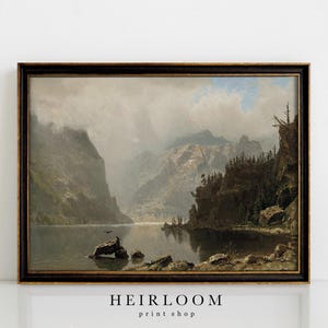 May include: A framed vintage landscape painting depicts a serene lake surrounded by towering mountains. The artwork features a dark wooden frame and a muted color palette of greens, grays, and browns, evoking a sense of tranquility. The text "HEIRLOOM print shop" is at the bottom.